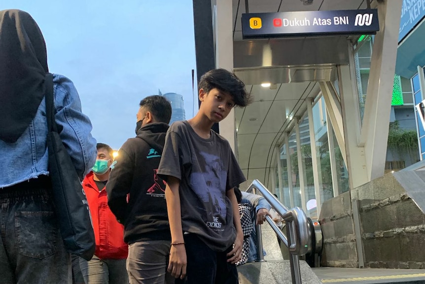 A teenage boy in a purple t-shirt looks at the camera against the background of the station entrance.