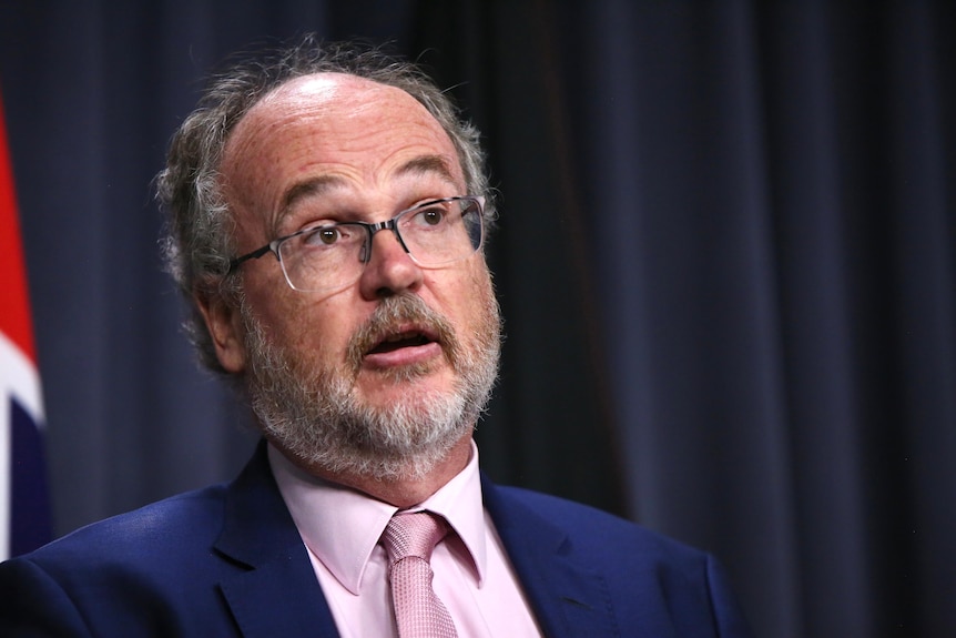 A man wearing a suit, tie and glasses answers questions at a press conference. 