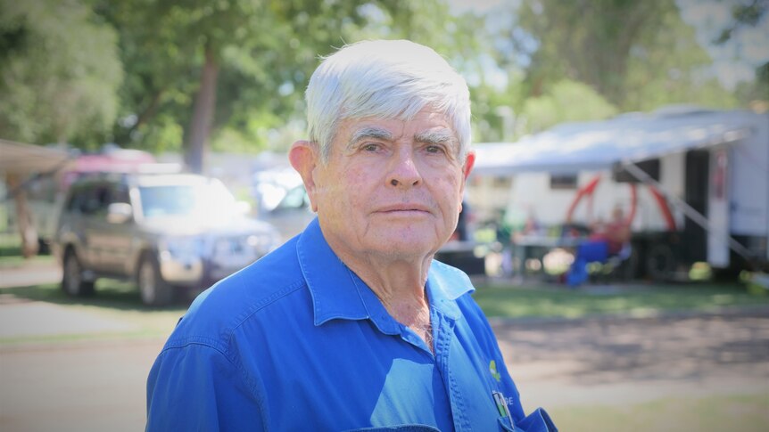 An older man with neat, white hair stands in a caravan park, looking solemn.