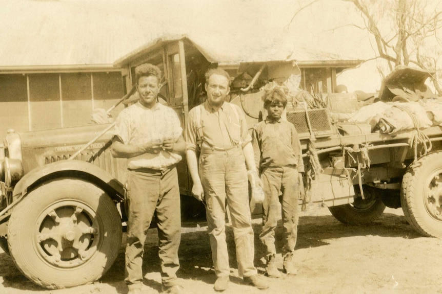 Two men and an Aboriginal boy stand in front of a 1920s/30s truck in a sepia-toned photo.
