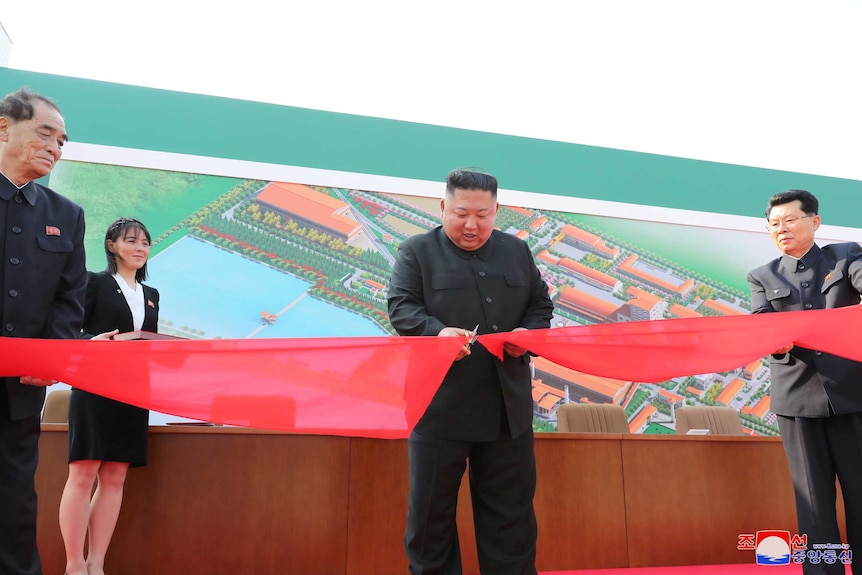 A stocky Asian man in a dark traditional suit cuts a red ribbon, watched by two men and one woman