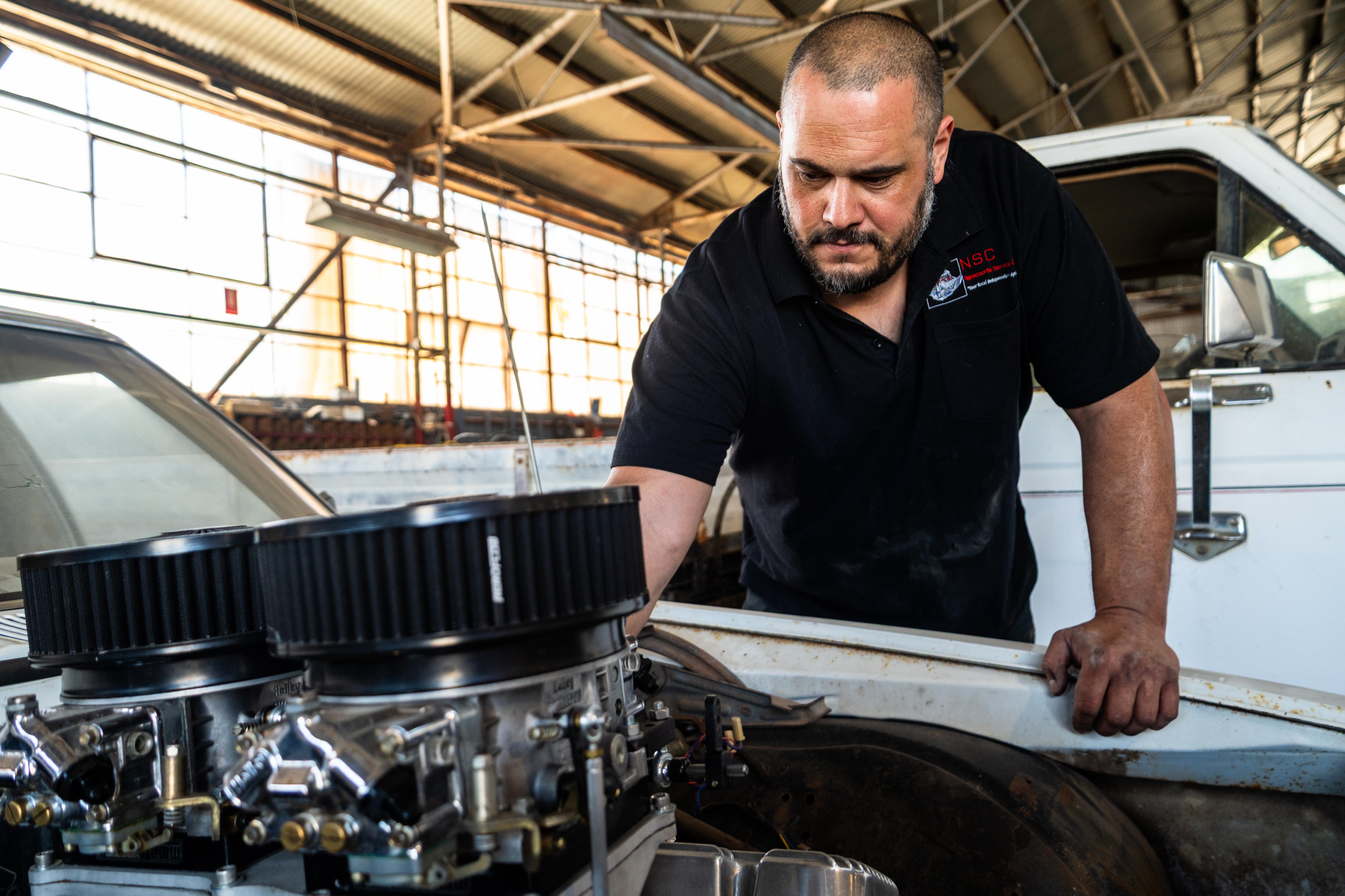 Um homem de camisa preta se apoia no capô de um carro em uma oficina