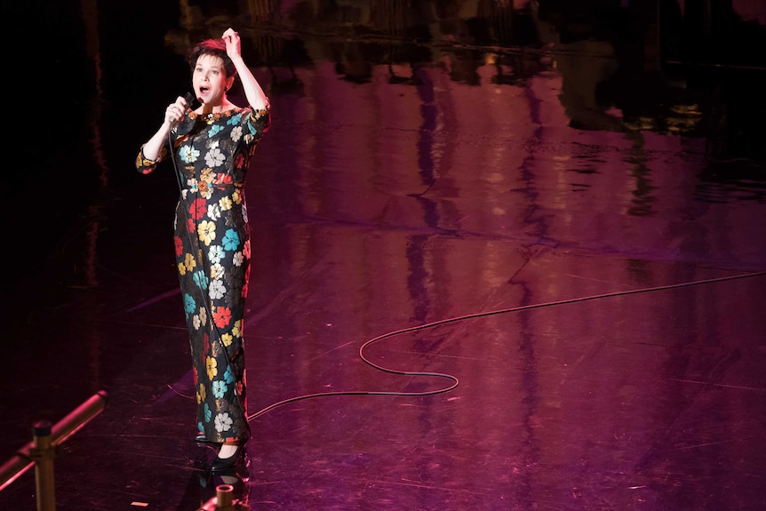 Renee Zellweger as Judy Garland, alone and singing on stage in a 70S flower jumpsuit