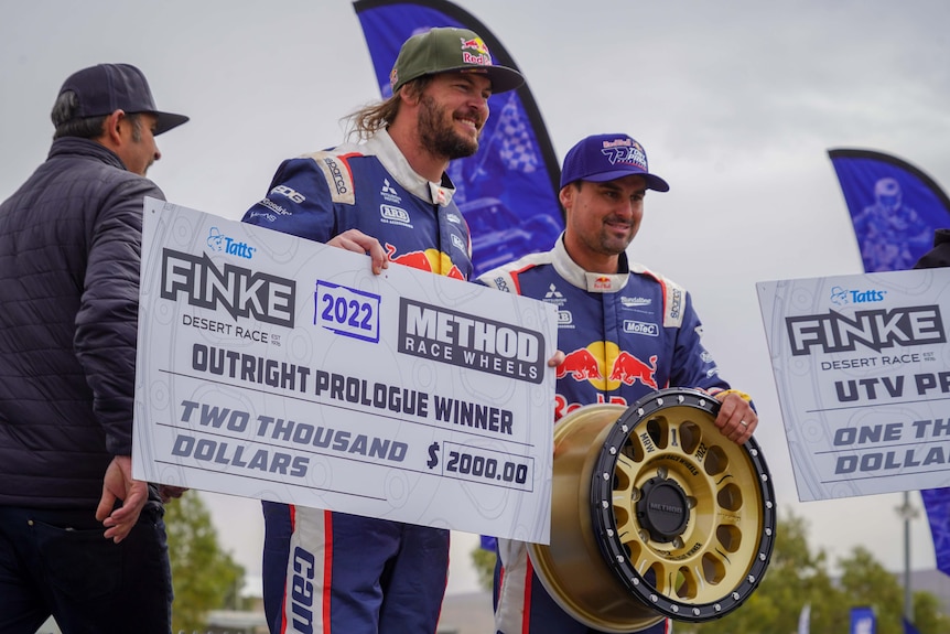 two men in red bull race suits smiling - one is holding a large cardboard cheque and the other is holding a golden tyre rim.