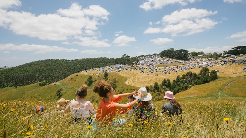 People sit in tall grass on a hillside with a festival set up in the background