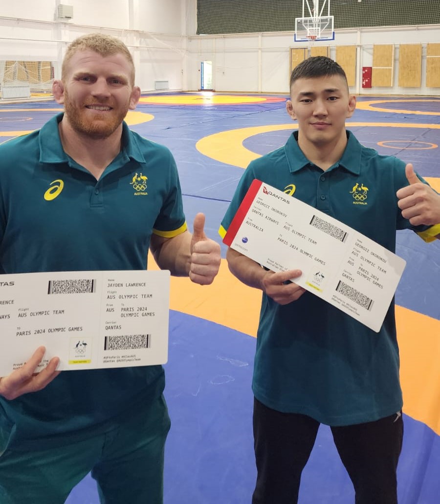 Australian wrestlers Jayden and Georgii hold their tickets to the Paris Olympic Games. They are smiling and giving thumbs up.