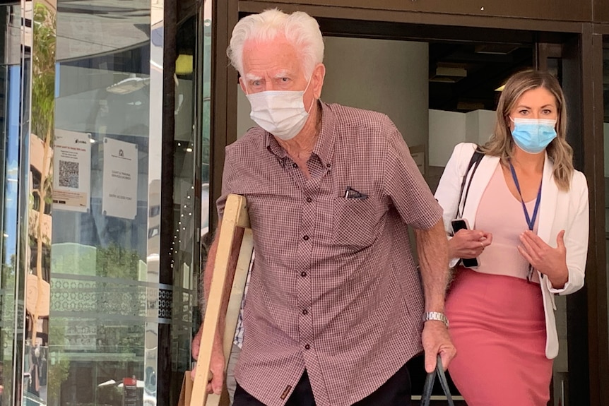 A mid-shot of an elderly man with white hair, a face mask and a burgundy shirt walking out of a court building.