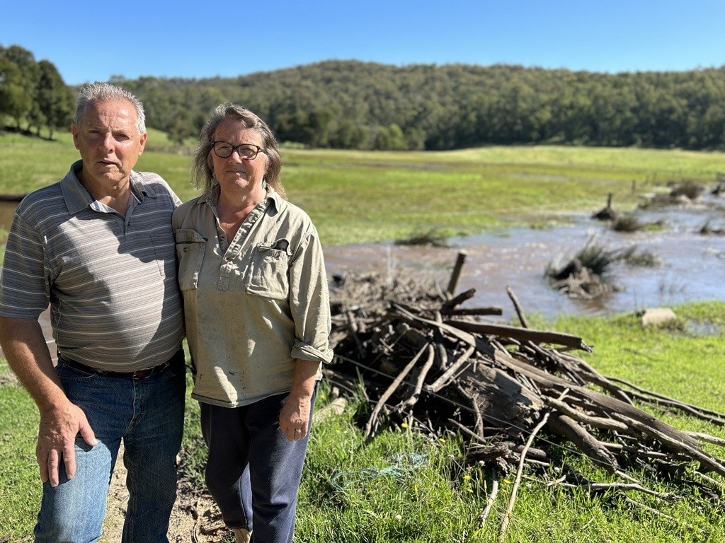 Damian and Angela Stock stand arm-in-arm in front of debris and flood water in their paddock