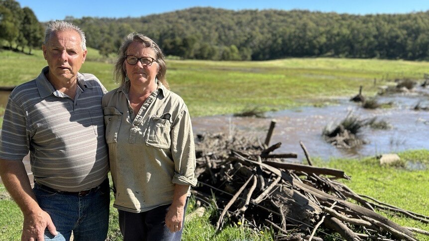 Damian and Angela Stock stand arm-in-arm in front of debris and flood water in their paddock