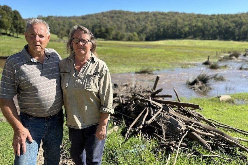 Damian and Angela Stock stand arm-in-arm in front of debris and flood water in their paddock