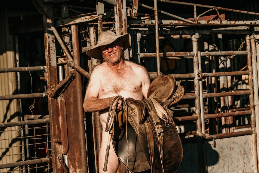 A naked farmer wearing a cowboy hat and holding a horse saddle