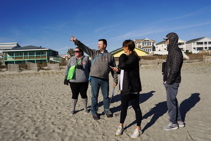 A group of four people point and look at the sky while standing on a beach in the middle of the day.