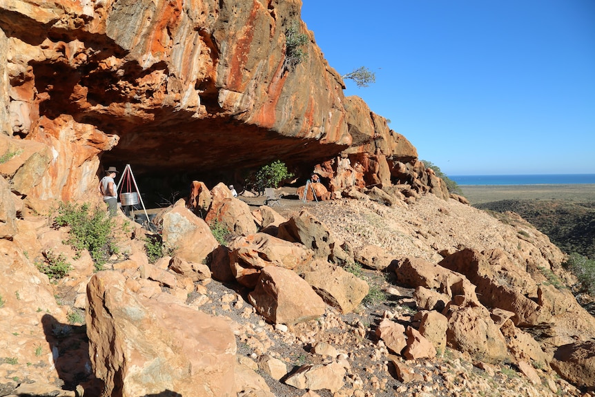 A red rock cave with research equipment in it. In the background there is a blue ocean and sky