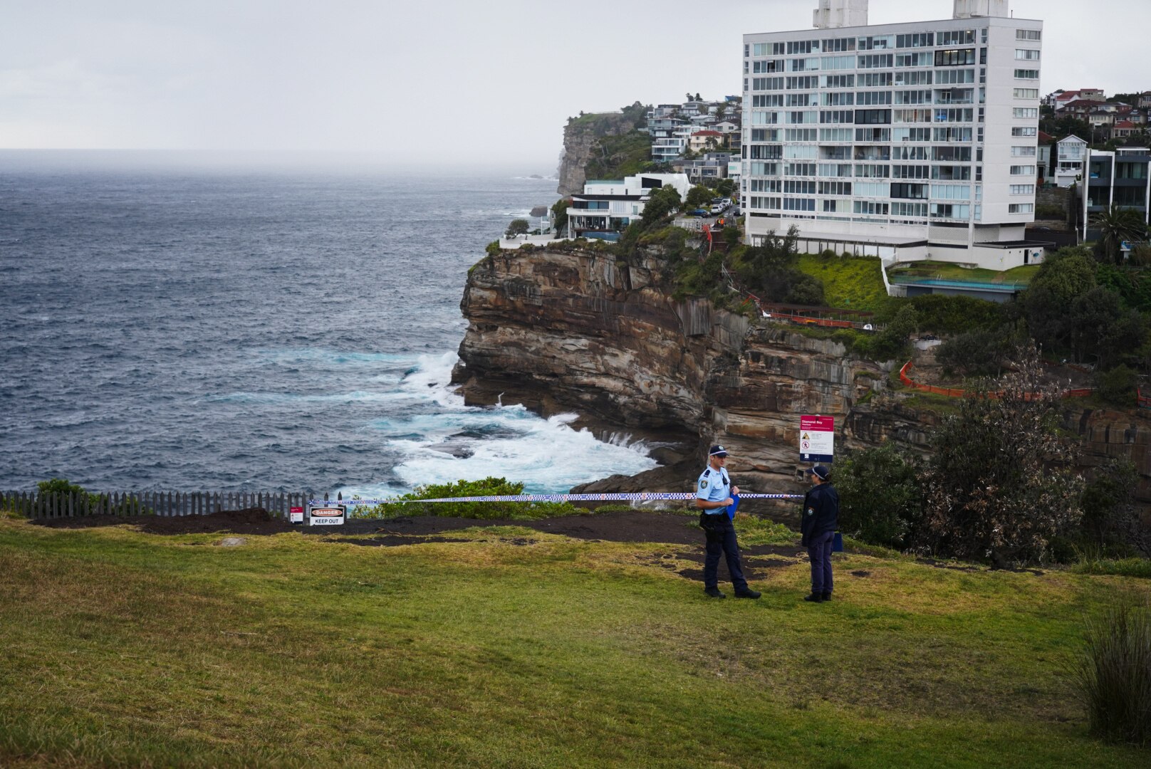 Police officers at Diamond Bay Reserve clifftop in Vaucluse with police tape behind them. Ocean and apratrtment block behind