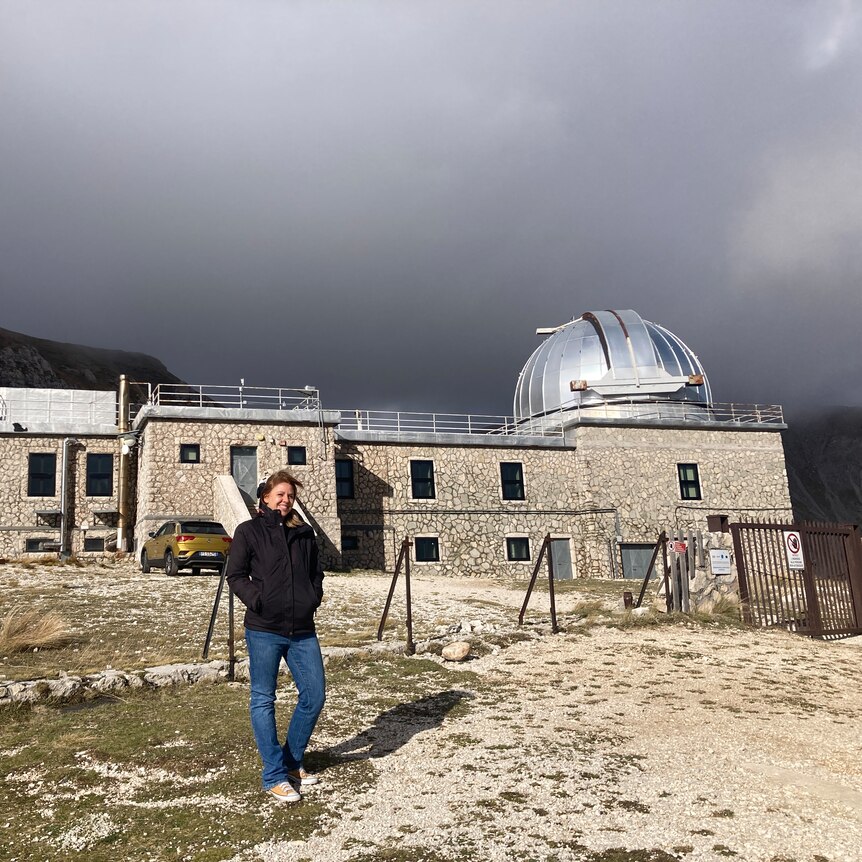 Dr Francesca Onori rugged up on a cold and windy mountaintop with a stone observatory building behind her
