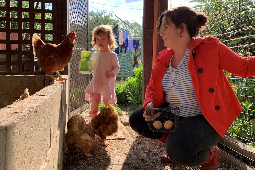 A woman in a red jacket and striped shirt crouches down inside a chicken coop with her young toddler collecting eggs.