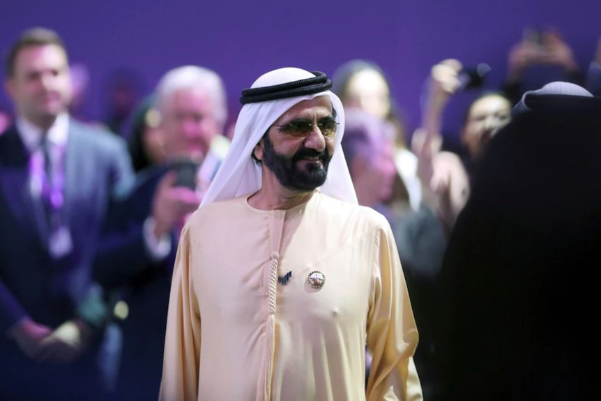 A man wearing a cream dress and white shall on his head walks through a crowd in a room lit with purple lights