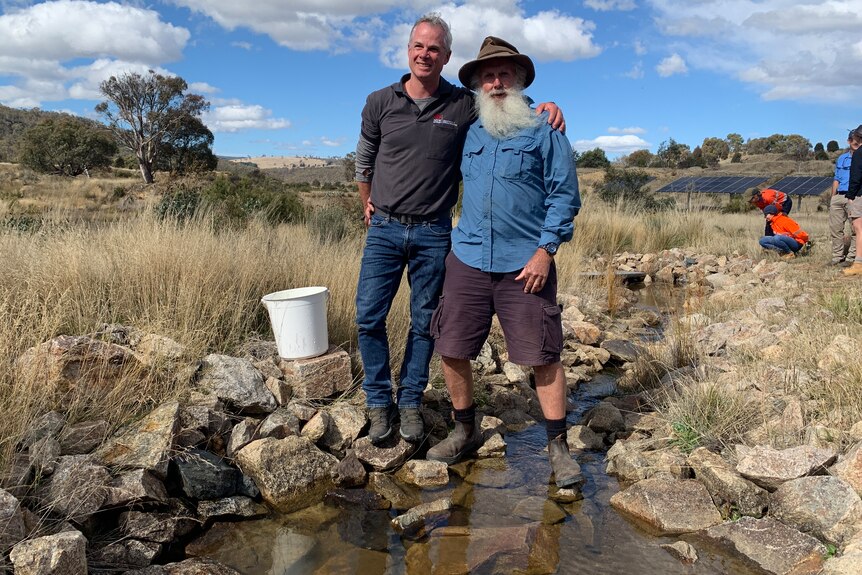 two men stand arm in arm by a river. They're smiling and holding a bucket where the fish were kept