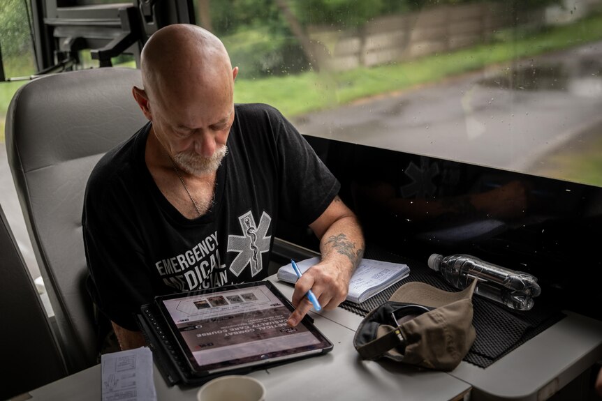 A bald man with grey-white facial hair sits at a desk inside a bus, looking at an ipad