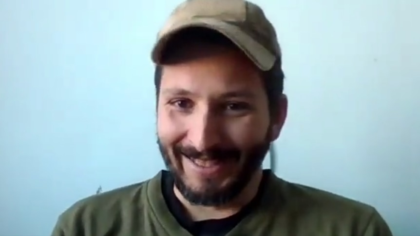 Wali smiles during a video link interview where he sits in front of a blue wall and wears a green cap.