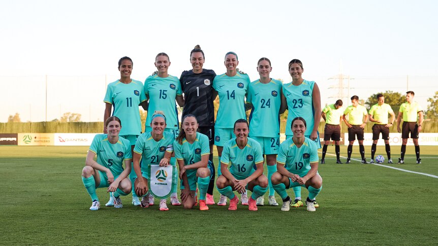 A women's soccer team wearing light blue poses for a team photo before a match with four referees in the background