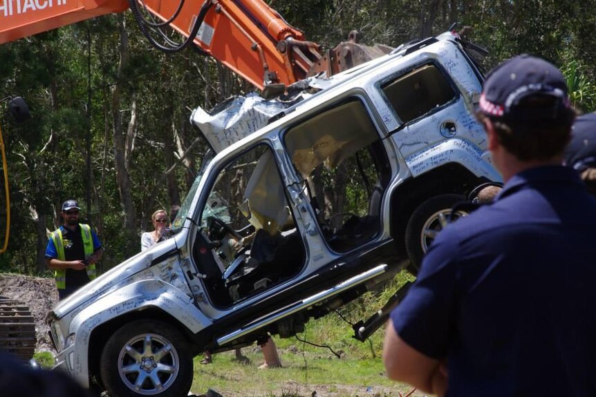 Jeep car held by a crane with doors taken off, writing all over it and people standing by watching it be destroyed.