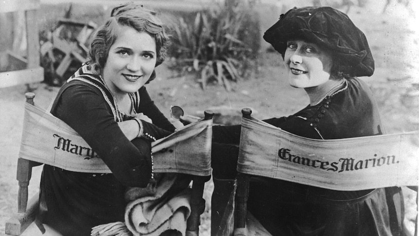 Black and white image of two women sitting in directors (deck) chairs and turning back to face the camera, smiling widely.