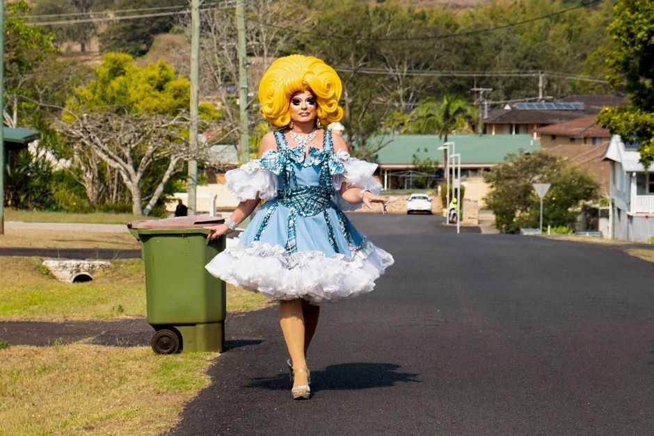 Drag queen in bright yellow wig and blue dress with wheelie bin