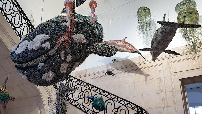 Marine sculptures from netting and ropes suspended from roof, staircase in background, large whale piece centre. 