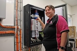 A woman in a pink and black polo shirt stands at a switchboard with a screwdriver in hand.