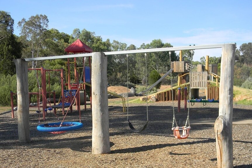 A playground partly made of timber with swings and a slide is located in a small clearing in the middle of a forest.