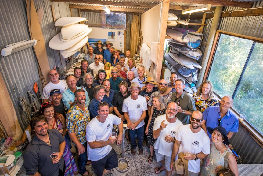 A crowd of people, smiling up at the camera, inside a shed lined with surfboards.