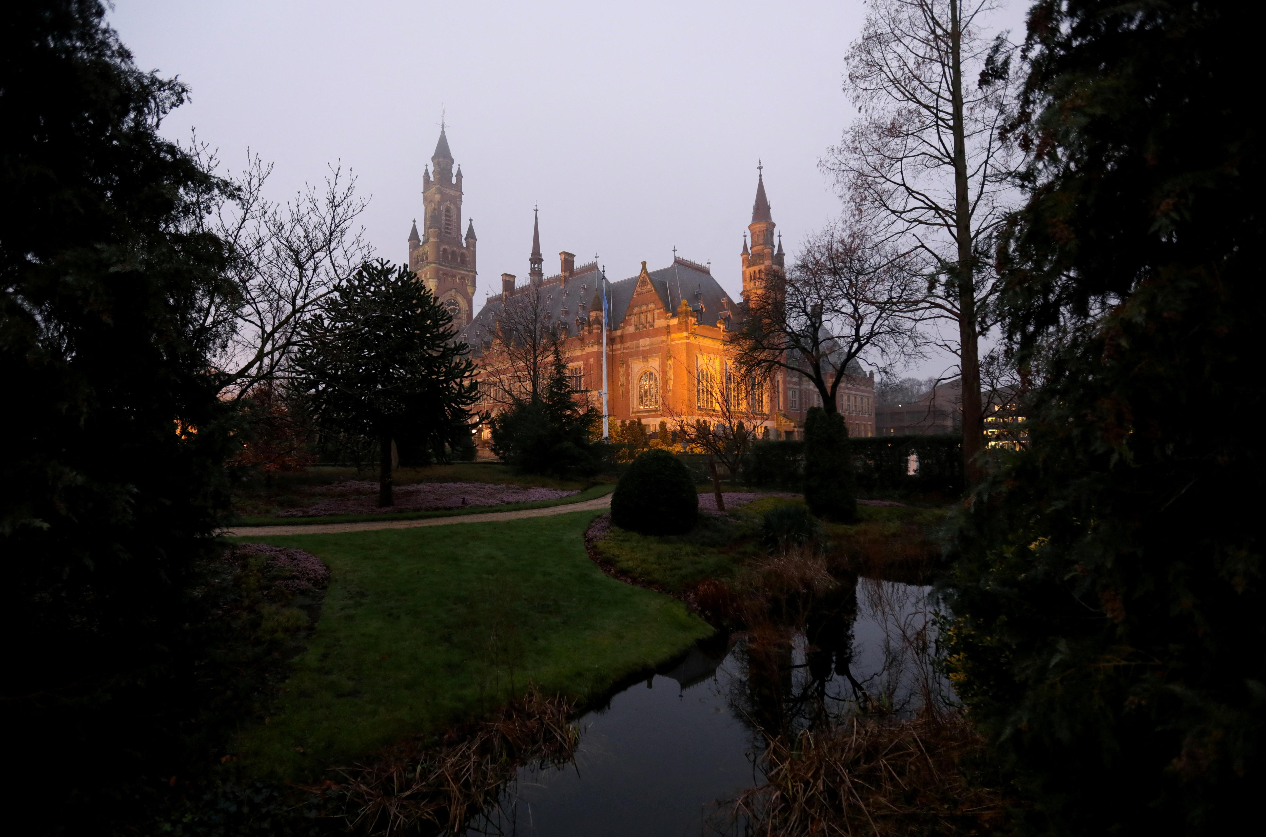 A building, illuminated by lights, is shown between trees with a grey sky