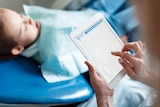 A little boy lies in a dental surgery, while a woman touches an screen.