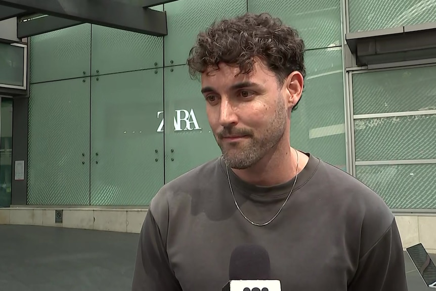 A young man with curly black hair wearing a grey t-short looks at a reporter during an interview in front of a shopping centr