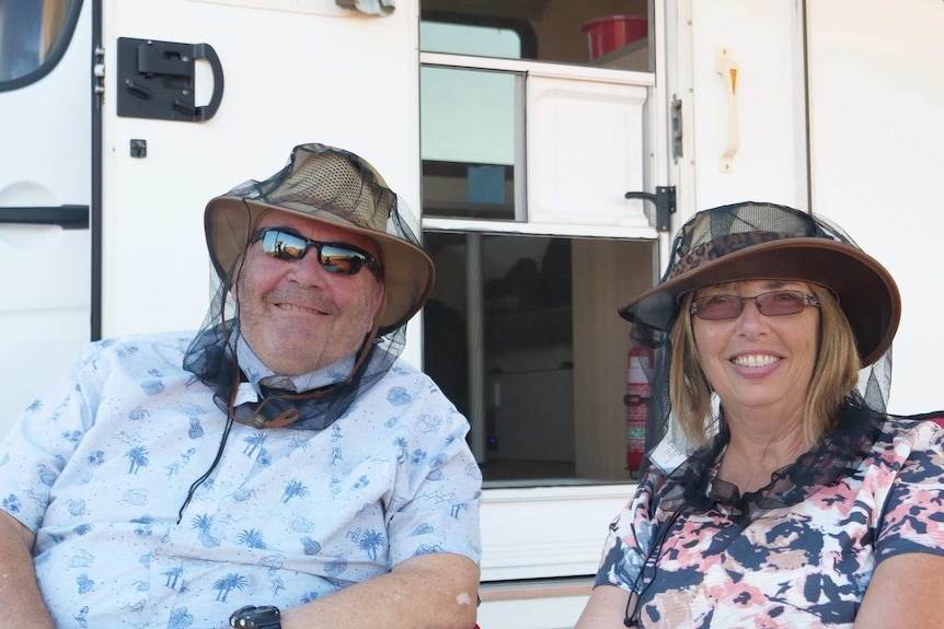 A male and female tourist sit in front of a caravan wearing sunglasses and hats with fly protectors.