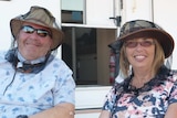 A male and female tourist sit in front of a caravan wearing sunglasses and hats with fly protectors.