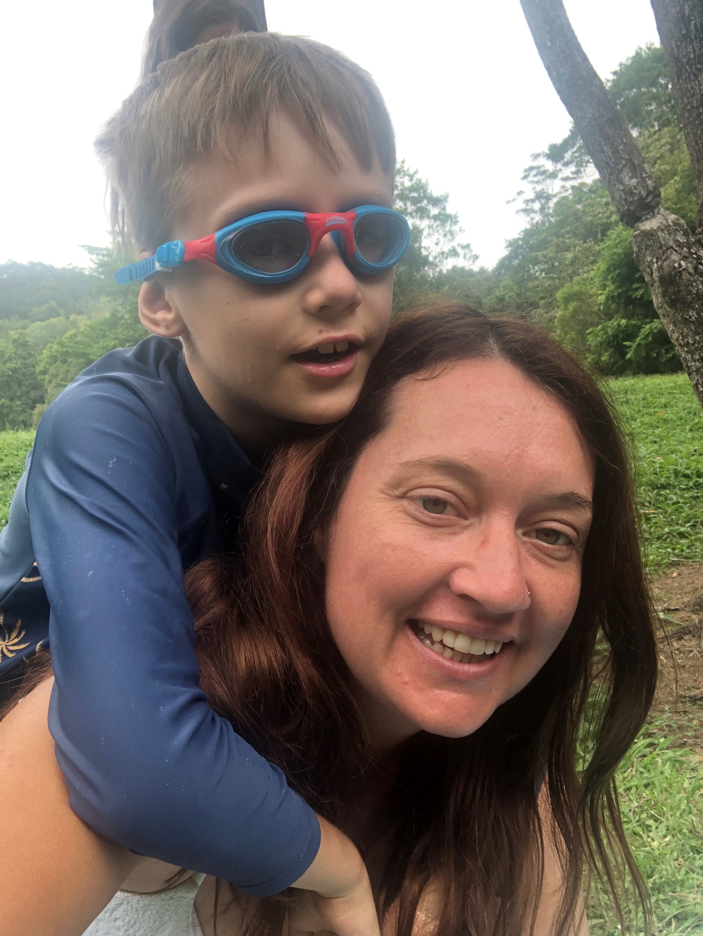 Young boy wearing goggles hangs over his Mum's back.