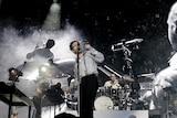 A group of musicians wearing black and white clothing play in front of a large black and white screen.