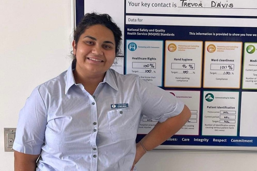 Chelsea standing in front of a board with writing on it, smiling, arm on hip, wearing nurses uniform.