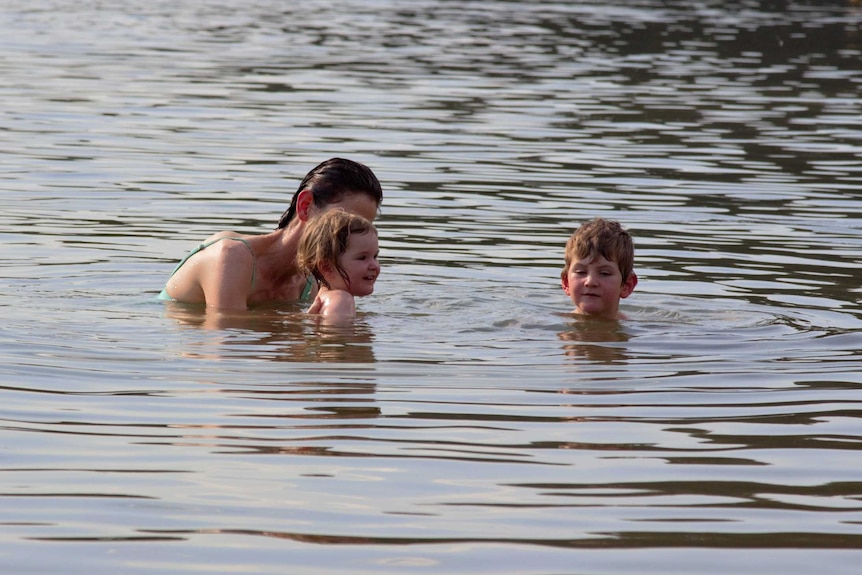 Woman holds young girl who is next to young boy, all swimming in water.