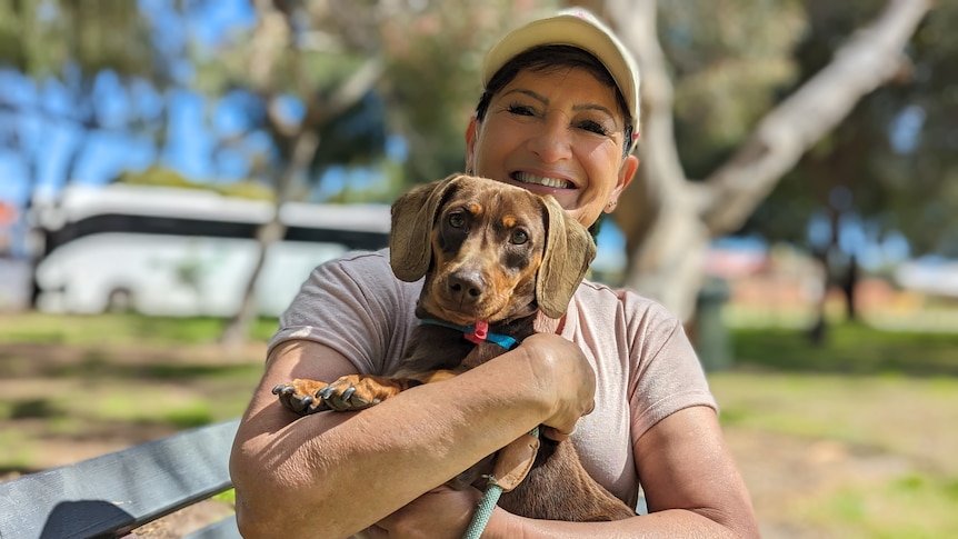 A smiling Marissa Ballard holds up dog Louis in a portrait taken outside in a park setting