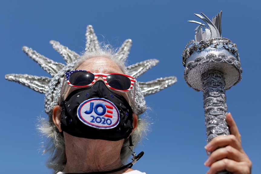 A grey-haired woman wearing a Joe face mask wears crown and hold baton like the Statue of Liberty.