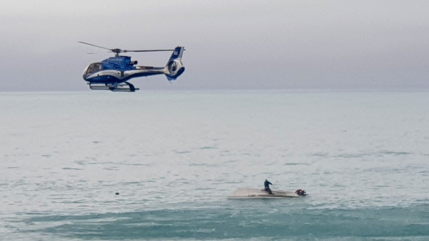 A helicopter flies overs an upturned boat with a survivor sitting on the hull 