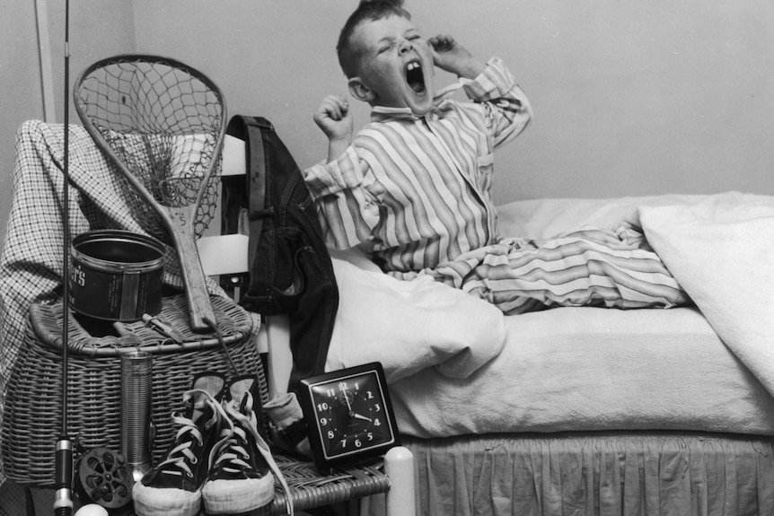 一张黑白照片中，一名刚睡醒的男孩床边放着他的球鞋和钓鱼装备。