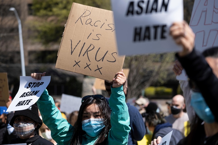 A young Asian woman in a face masks holds up different signs with her hands in daytime rally.