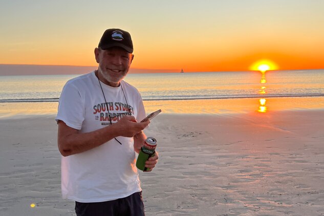 A smiling older man holding a phone and a can in a stubby holder as the sun sets over the beach behind him.