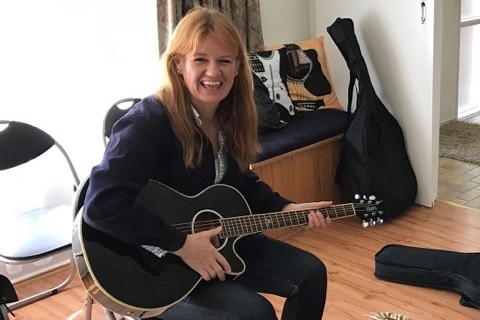 Jodi Ball sitting in the middle of a room on a chair playing guitar and smiling at the camera.