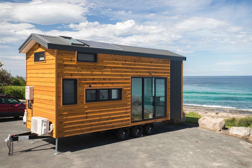 A tiny home parked by the ocean. It is one and a half storeys high and clad in light wood. It is on a trailer that can be towed