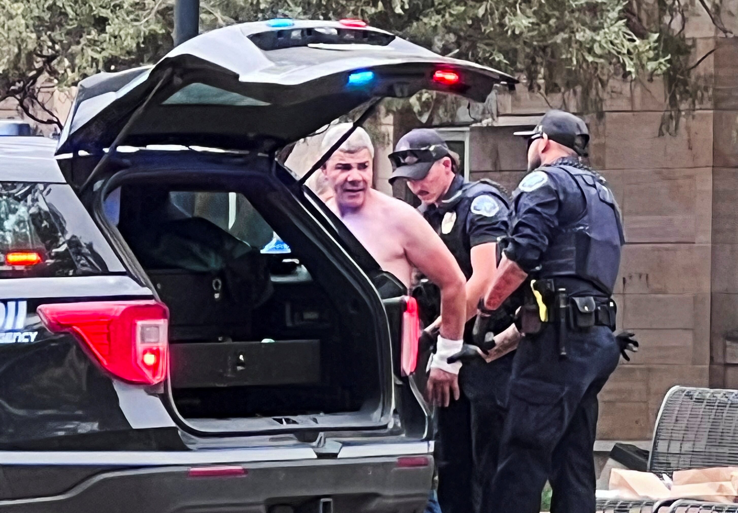 A greying, shirtless man with his arms being pulled behind his back by two police officers in black clothing next to a car